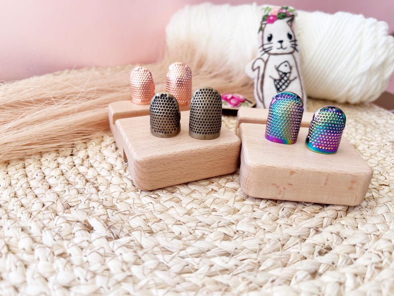 May include: A collection of thimbles in various colors and finishes, including copper, bronze, and iridescent rainbow. The thimbles are displayed on small wooden blocks, with a white yarn ball and a cat illustration in the background.