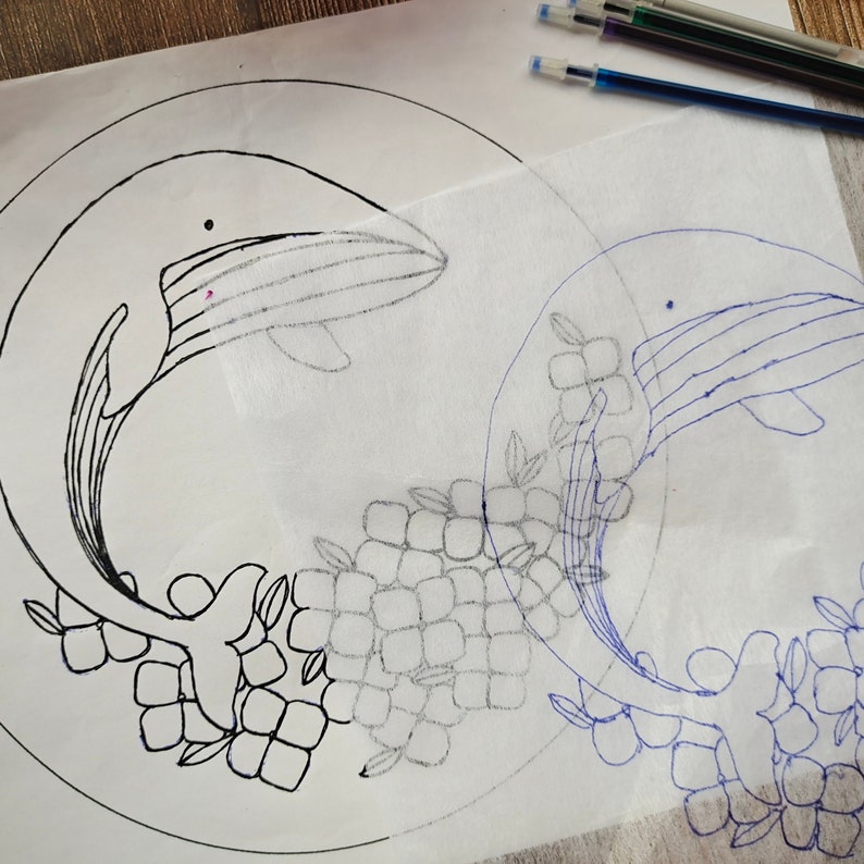 Pu&ograve; includere: Un disegno in bianco e nero di una balena con fiori su sfondo bianco. Un tracciato blu della balena e dei fiori &egrave; visibile su un foglio trasparente posto sopra il disegno.