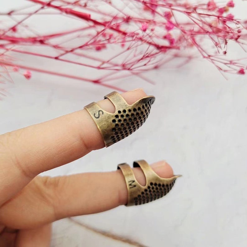 May include: Two gold-toned thimbles for sewing, one marked "S" and the other marked "M".