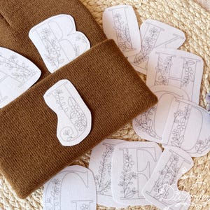 May include: A brown knit beanie with several white floral letter patches. The patches feature the letters A, B, E, F, G, H, I, J, N, and O, each decorated with delicate floral designs. The items are arranged on a woven surface.