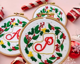 May include: Three embroidered Christmas ornaments with a holly wreath design and a red letter in the center of each. The letters are A, P, and J. The ornaments are on a pink background.