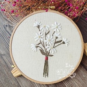 Op de afbeelding: Een witte borduurring met een boeket witte bloemen met groene stelen en bladeren. De bloemen zijn met een rood lint aan elkaar gebonden.