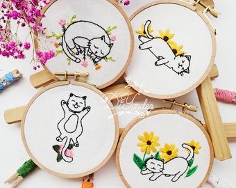 May include: Four embroidery hoops with white fabric and black embroidery of cats. The cats are in different poses, including sleeping, sitting, and lying down. The hoops are on a wooden surface.