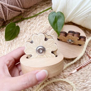 Puede incluir: Un devanador de hilo de madera con un diseño de margarita, sostenido en una mano. El devanador es de color marrón claro con un centro plateado. Se ve un segundo devanador más pequeño, junto con hilo de color crema y beige, y unas tijeras de oro rosa. El fondo es una estera tejida.