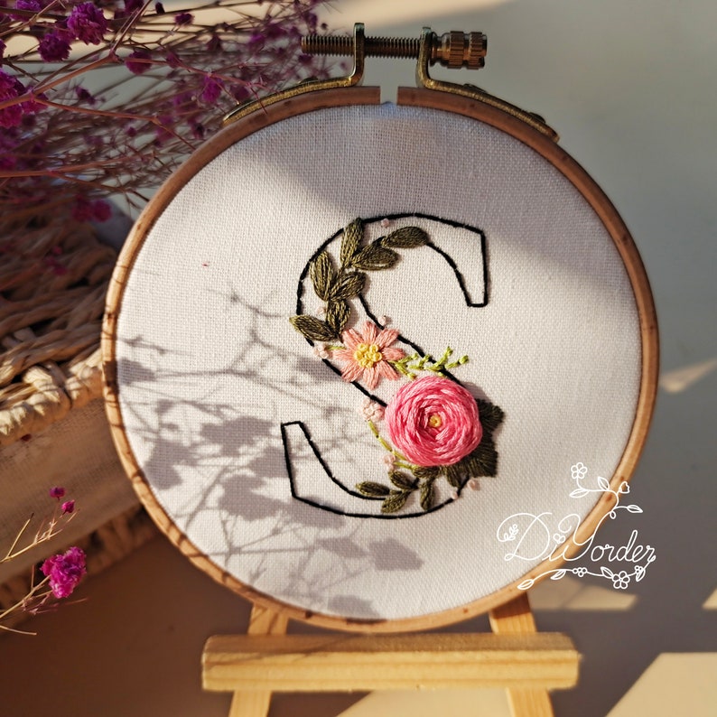 May include: Embroidered floral letter S in a white hoop with a wooden stand. The letter is surrounded by pink and green flowers.
