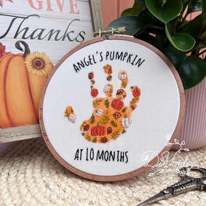 Könnte beinhalten: Gesticktes Stickrahmen-Kunstwerk mit einem Handabdruck-Design, gefüllt mit Kürbissen und Herbstlaub. Der Text "Angel's Pumpkin at 10 Months" ist oberhalb und unterhalb des Handabdrucks gestickt. Der Rahmen steht vor einem Hintergrund mit einem Kürbis und dem Schriftzug "Give Thanks".