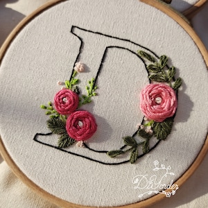 May include: Embroidered hoop art featuring the letter D with pink flowers and green leaves. The letter is outlined in black thread.