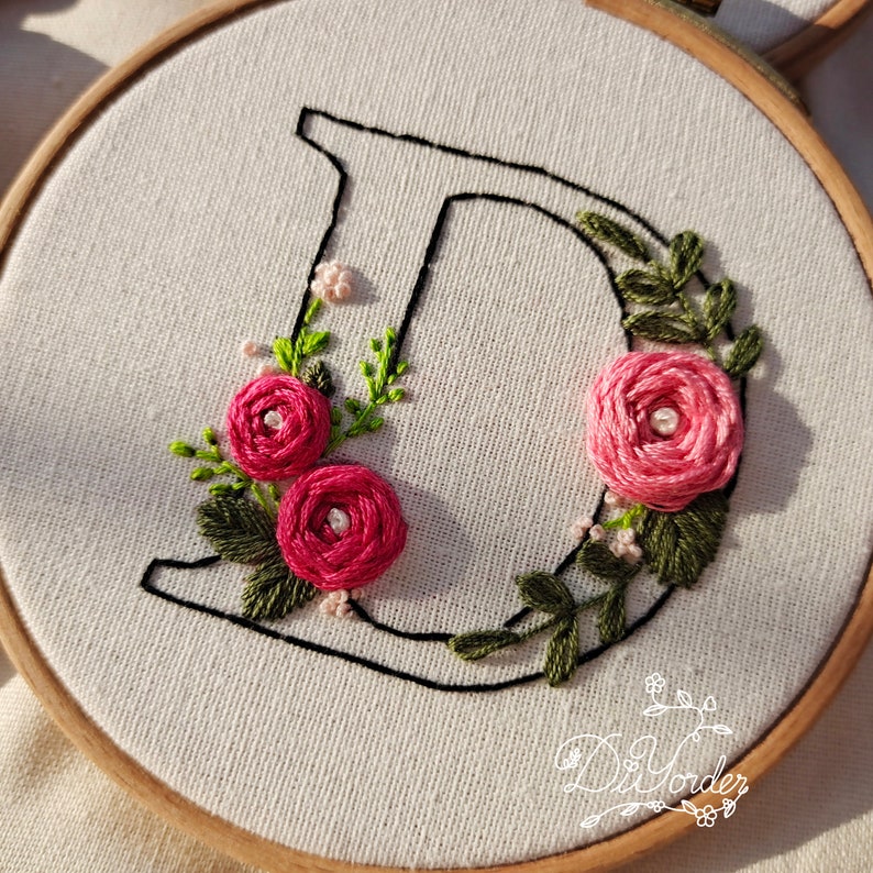 May include: Embroidered hoop art featuring the letter D with pink flowers and green leaves. The letter is outlined in black thread.