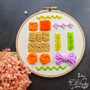 Könnte beinhalten: Nahaufnahme eines runden Stickrahmens mit verschiedenen bunten Stickereien. Die Stiche umfassen Zickzack, einen flauschigen Bommel, ein Quadrat, einen gewebten Stich, einen Zopf, ein Blatt, eine Blume und eine Schleife. Der Rahmen ist aus Holz und hat einen weißen Stoffhintergrund.