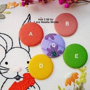 May include: Five colorful needle minders, labeled A through E, are displayed on a white fabric with embroidery. The minders are round and made of fabric in orange, pink, purple floral, yellow, and green. The text "Add 2.99 for 1 pcs Needle Minder" is also visible.