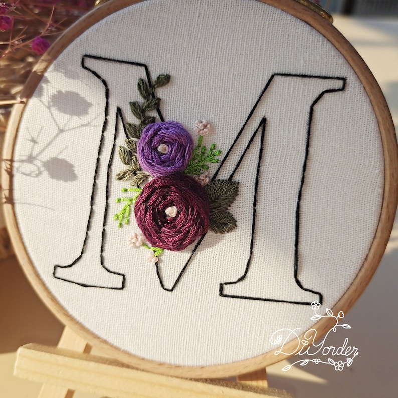 May include: Embroidered letter "M" with purple and burgundy flowers on a white fabric background. The embroidery is in a hoop with a wooden frame.