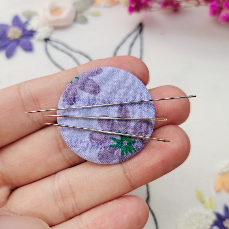 May include: Close-up of three sewing needles resting on a small, round, lavender fabric piece with a floral pattern. The needles are silver and gold in color. The fabric has purple flowers and green leaves.