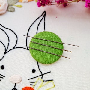 May include: Three silver embroidery needles are arranged on a green circular pincushion. The pincushion is on a white fabric background with a black embroidered design of a rabbit.