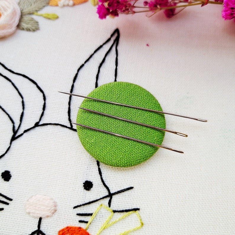 May include: Three silver embroidery needles are arranged on a green circular pincushion. The pincushion is on a white fabric background with a black embroidered design of a rabbit.