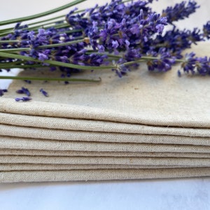 Pu&ograve; includere: Un mucchio di otto strofinacci di lino naturale, piegati con cura. Gli strofinacci sono di colore beige chiaro e hanno una texture sottile. Un mazzo di fiori di lavanda riposa sopra gli strofinacci.