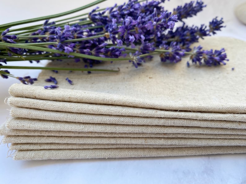 Pu&ograve; includere: Un mucchio di otto strofinacci di lino naturale, piegati con cura. Gli strofinacci sono di colore beige chiaro e hanno una texture sottile. Un mazzo di fiori di lavanda riposa sopra gli strofinacci.