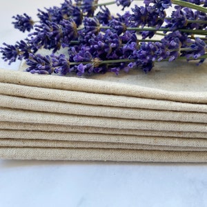 Pu&ograve; includere: Un mucchio di otto tovaglioli di lino naturale, piegati e disposti ordinatamente. I tovaglioli sono di colore beige chiaro e hanno una texture sottile. Un rametto di fiori di lavanda &egrave; posto sopra i tovaglioli.