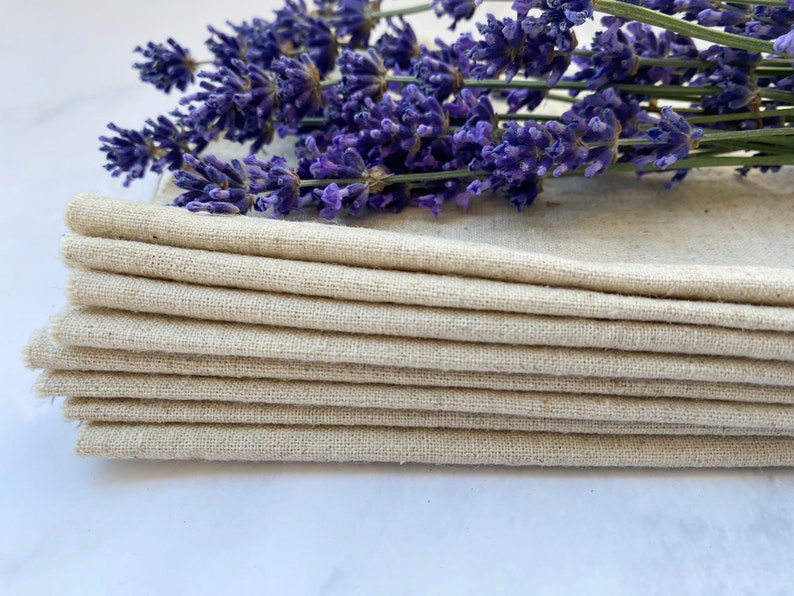 Pu&ograve; includere: Un mucchio di otto tovaglioli di lino naturale, piegati e disposti ordinatamente. I tovaglioli sono di colore beige chiaro e hanno una texture sottile. Un rametto di fiori di lavanda &egrave; posto sopra i tovaglioli.