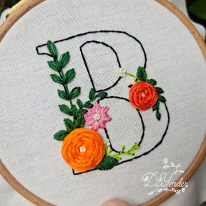 May include: Embroidered letter B with floral embellishments. The letter is outlined in black thread and surrounded by green leaves, orange and pink flowers.
