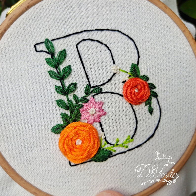 May include: Embroidered letter B with floral embellishments. The letter is outlined in black thread and surrounded by green leaves, orange and pink flowers.