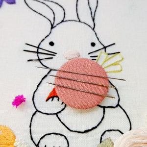 May include: Embroidery project featuring a bunny design with a pink circle and three needles. The bunny is outlined in black thread, with a pink nose and orange carrot. Colorful floral embroidery accents the design.