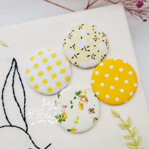May include: Four yellow fabric covered buttons with different patterns. The buttons are arranged in a circle on a white fabric background. The patterns include a floral design, polka dots, gingham, and a small floral print.