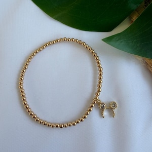 May include: Gold beaded bracelet with a charm featuring two clear, round gemstones. The bracelet is made of small, round gold beads. The charm is in the shape of a stylized head with two round gemstones. The bracelet is on a white surface.