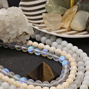 May include: A collection of gemstone bead necklaces in shades of white, cream, and iridescent. A small, brown pyramid-shaped stone sits among the necklaces. A tiered serving tray holds various polished stones and crystals. The background is dark.