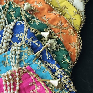 Może przedstawiać: Kolekcja kolorowych, haftowanych torebek z tkaniny w odcieniach różu, niebieskiego, zielonego, pomarańczowego, żółtego i białego. Każda torebka ma złote haftowane kwiaty, obszycia z koralików i ozdobne frędzle.