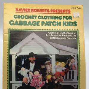 May include: A vintage book cover titled "CROCHET CLOTHING FOR CABBAGE PATCH KIDS" by Xavier Roberts. The cover features a photo of a man holding several Cabbage Patch Kids dolls, with a white gazebo in the background. The book is yellow with green and red accents.