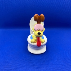 May include: A yellow Garfield figurine sitting on a white toilet with a blue rim. Garfield is wearing a brown collar and has a red tongue sticking out.