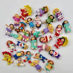 May include: A collection of small Cabbage Patch Kids plastic figurines featuring various characters in diverse outfits, poses, and accessories, including umbrellas, sports gear, and heart-shaped signs with the text I LOVE YOU printed on them.