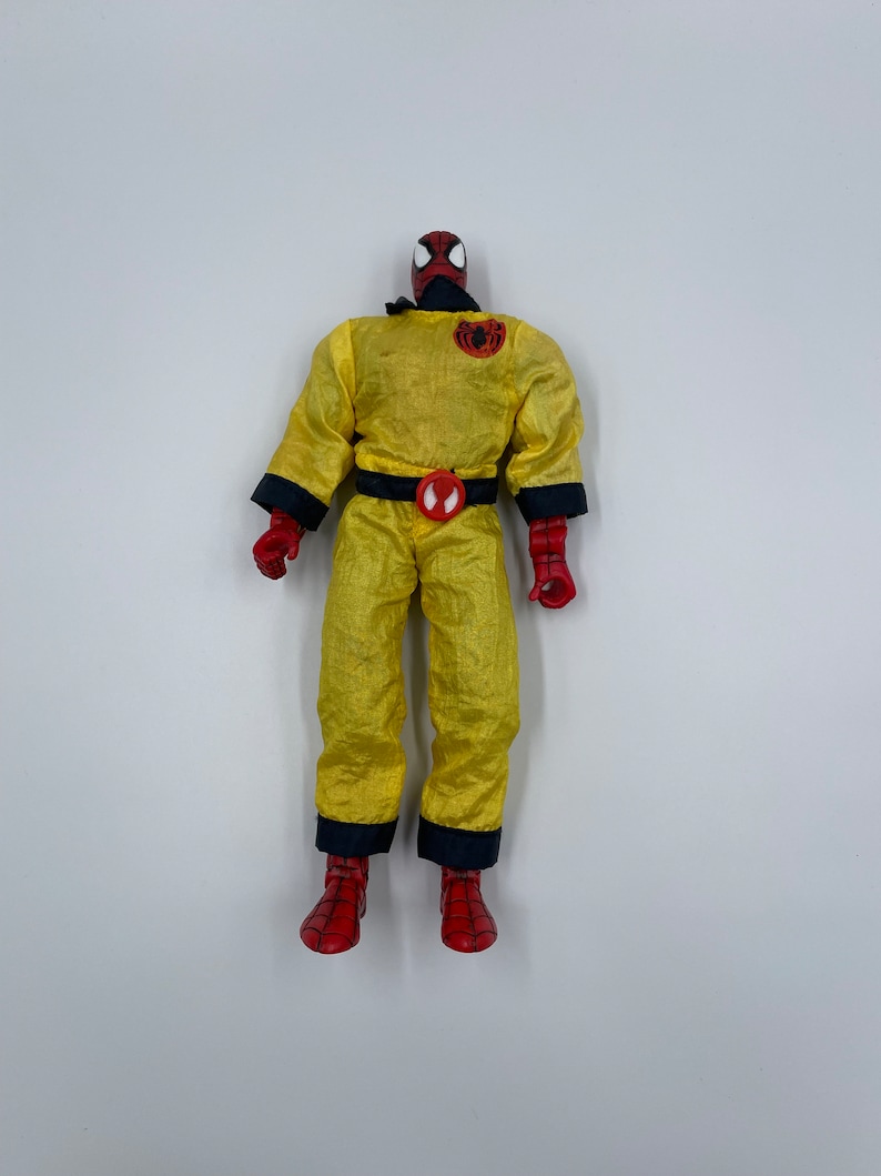 Puede incluir: Una figura de acci&oacute;n de Spider-Man de color amarillo y rojo que lleva un mono amarillo con un cintur&oacute;n negro y un logotipo de ara&ntilde;a en el pecho. La figura tiene la cabeza roja y los ojos negros.