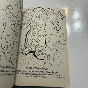 May include: A black and white maze with a winged warrior design. The maze has a start point and a finish point. The text "26. WINGED WARRIOR" is at the top of the maze. The text "Goldar is going to get Red Ranger, Black Ranger, and Yellow Ranger unless you find the path that leads them through the maze." is below the maze.