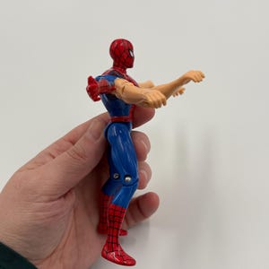 May include: A Spider-Man action figure in a dynamic pose. The figure is predominantly blue and red, with a red mask and web detailing. The figure's arms are outstretched, and the figure is held by a hand.