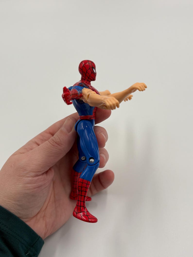 May include: A Spider-Man action figure in a dynamic pose. The figure is predominantly blue and red, with a red mask and web detailing. The figure's arms are outstretched, and the figure is held by a hand.