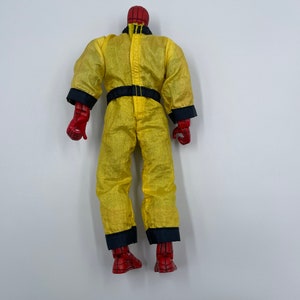Puede incluir: Una figura de acci&oacute;n roja y azul que lleva un mono amarillo con ribetes negros. La figura est&aacute; de pie sobre un fondo blanco.