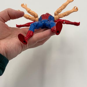 May include: A Spider-Man action figure with multiple arms extending from its back. The figure is in a lying position, with red and blue colors. The figure is held in a hand against a white background.