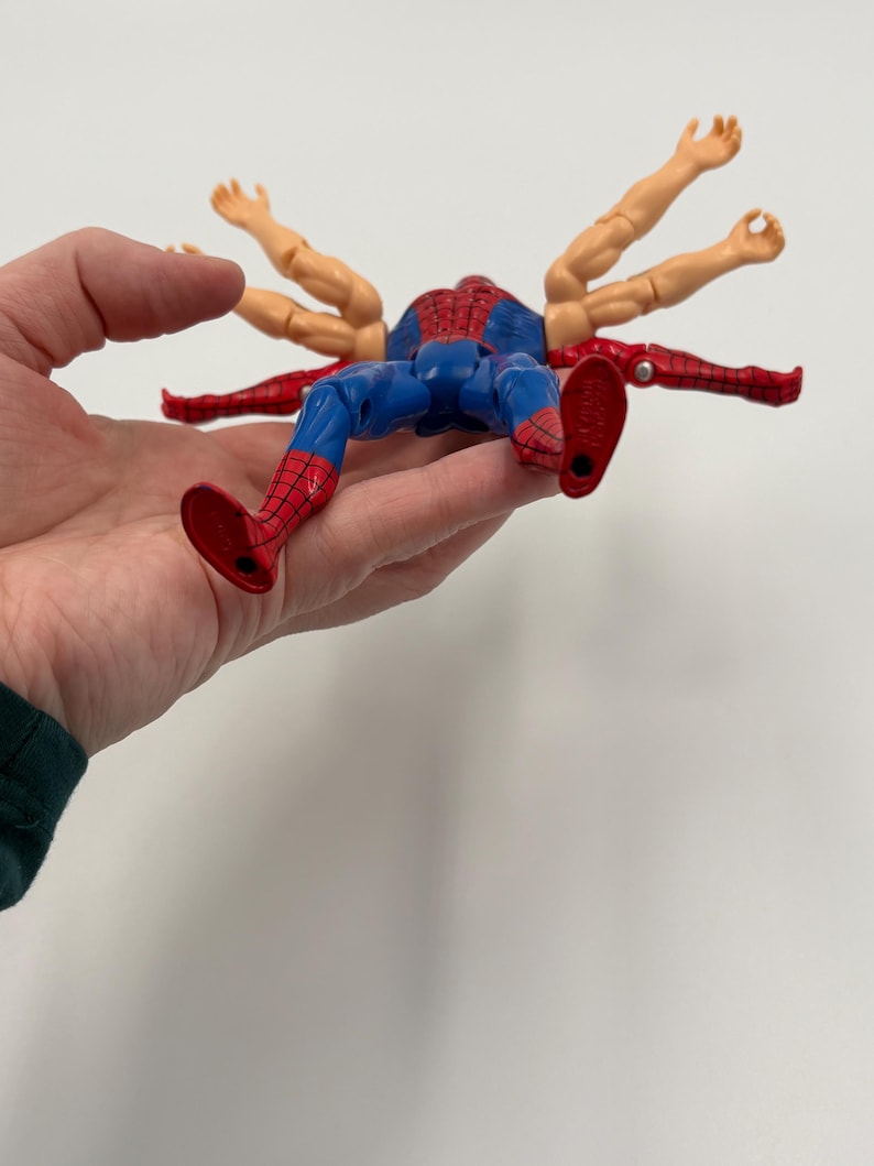 May include: A Spider-Man action figure with multiple arms extending from its back. The figure is in a lying position, with red and blue colors. The figure is held in a hand against a white background.
