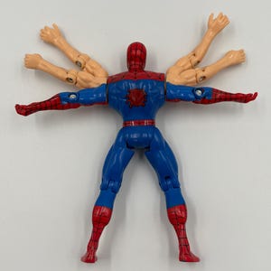 May include: A blue, red, and tan Spider-Man action figure with multiple arms. The figure has a red mask and spider emblem on the chest. The legs and arms are articulated.