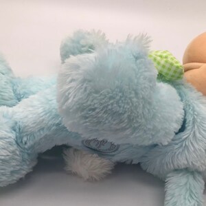 K&ouml;nnte beinhalten: Ein blauer Pl&uuml;schhase mit einer gr&uuml;n-wei&szlig; karierten Schleife. Der Hase liegt auf der Seite mit dem Kopf nach rechts gedreht. Der Hase ist aus weichem, flauschigem Material.