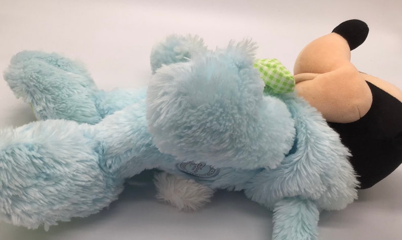 K&ouml;nnte beinhalten: Ein blauer Pl&uuml;schhase mit einer gr&uuml;n-wei&szlig; karierten Schleife. Der Hase liegt auf der Seite mit dem Kopf nach rechts gedreht. Der Hase ist aus weichem, flauschigem Material.