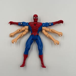 May include: A Spider-Man action figure with six arms, in a classic red and blue costume. The figure has a detailed design, with a spider emblem on the chest and red boots. The figure is posed with arms outstretched.