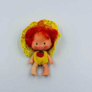 May include: A small doll with bright orange hair, wearing a yellow hat with green polka dots and a yellow outfit featuring a red apple graphic on the chest. The doll has a light skin tone, freckles, and green eyes, with one leg lighter than the other.