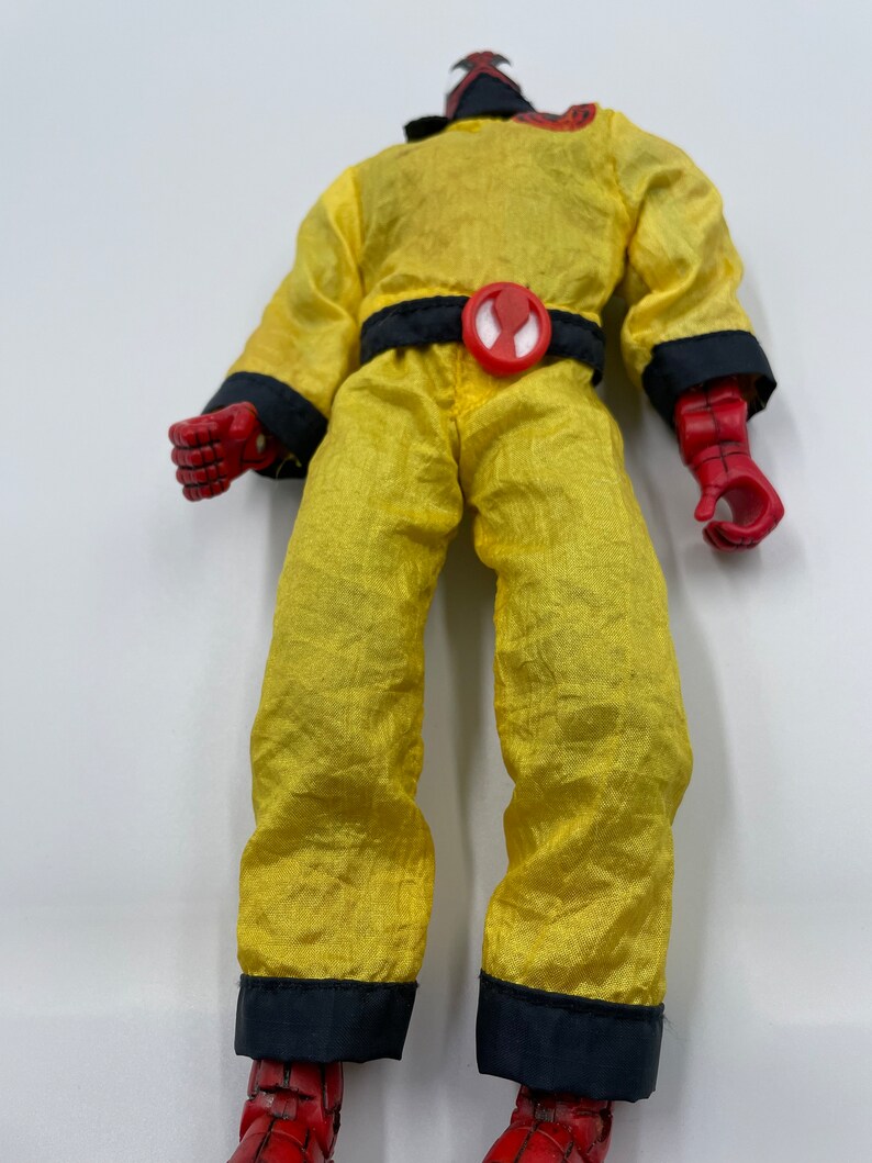 Puede incluir: Un mono amarillo con ribetes negros y un s&iacute;mbolo de ara&ntilde;a roja en el cintur&oacute;n. La figura lleva una m&aacute;scara y guantes rojos.