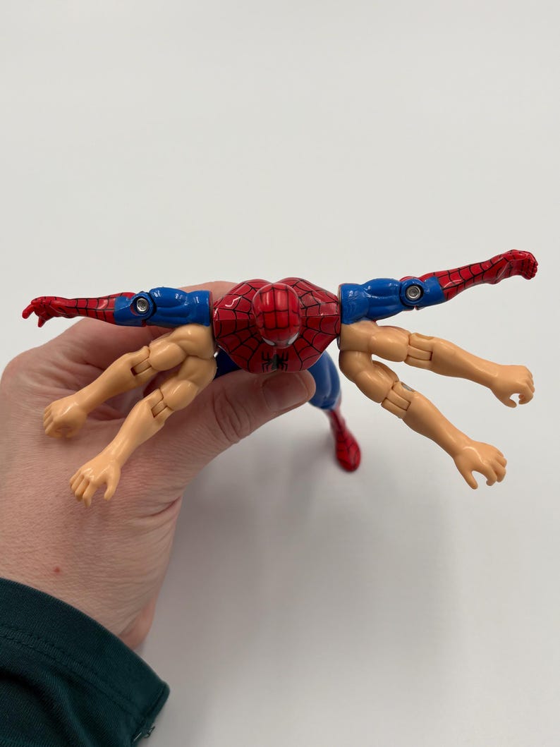May include: A red, blue, and tan Spider-Man action figure with multiple arms. The figure has a red spider-web design on the chest and head. The arms are in various positions, with some extended and others bent.
