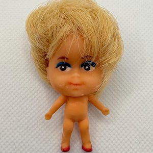 May include: A small plastic doll with blonde hair, blue eyeshadow, and red shoes. The doll is nude and has a simple facial expression.