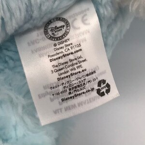 K&ouml;nnte beinhalten: Ein wei&szlig;es Etikett mit schwarzem Text, "GENUINE ORIGINAL DISNEY STORE AUTHENTIC" mit einem Disney Store Logo. Das Etikett enth&auml;lt auch die Adresse und die Website des Disney Stores, "DisneyStore.com", und den Text "100%" mit einem Recycling-Symbol.