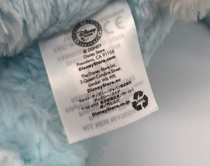 K&ouml;nnte beinhalten: Ein wei&szlig;es Etikett mit schwarzem Text, "GENUINE ORIGINAL DISNEY STORE AUTHENTIC" mit einem Disney Store Logo. Das Etikett enth&auml;lt auch die Adresse und die Website des Disney Stores, "DisneyStore.com", und den Text "100%" mit einem Recycling-Symbol.