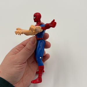 May include: A Spider-Man action figure in a dynamic pose. The figure features a red and blue costume with a red mask. The figure's arms are extended, with one hand in a thumbs-up gesture. The figure is held against a white background.